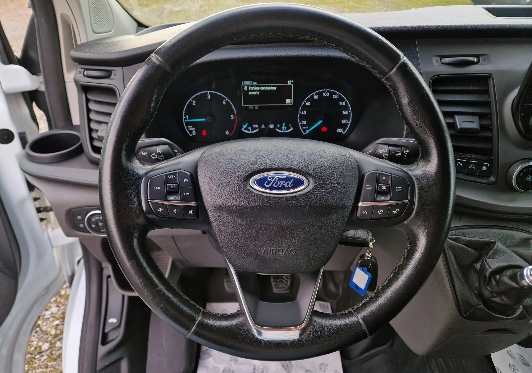 Vue intérieure centrée sur le volant noir du Ford Transit Custom Fourgon blanc, avec tableau de bord et levier de vitesses manuel visibles.
