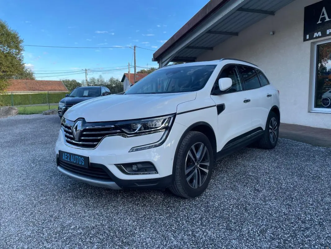 Renault Koleos blanc vu de 3/4 avant droit, avec calandre chromée et jantes alliage foncé, stationné devant un bâtiment.