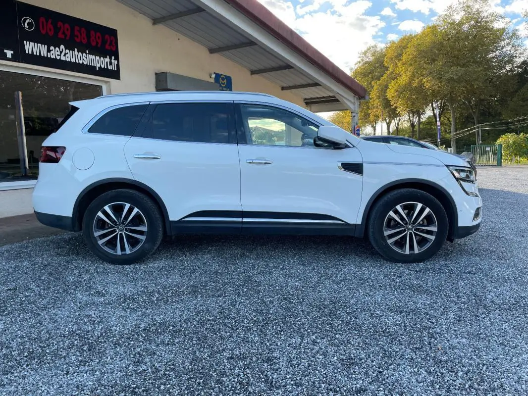 Renault Koleos blanc vu de profil côté gauche, avec jantes alliage et vitres teintées sur parking extérieur.