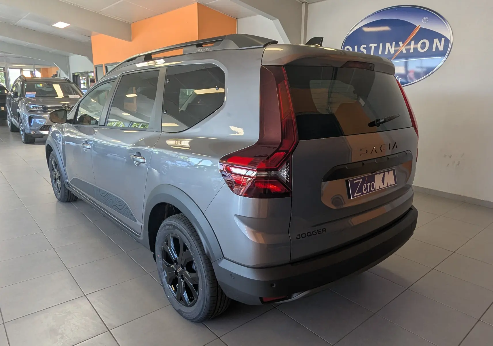 Vue 3/4 arrière droite du Dacia Jogger gris schiste métal avec jantes noires et toit équipé de barres de toit en showroom.