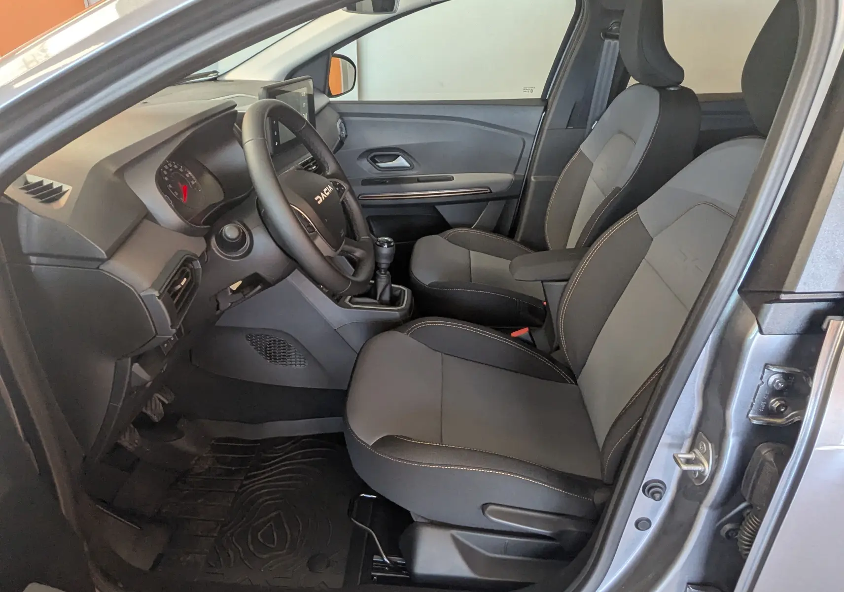 Intérieur avant droit du Dacia Jogger gris schiste, sièges tissu noir avec surpiqûres, volant multifonction et boîte manuelle.