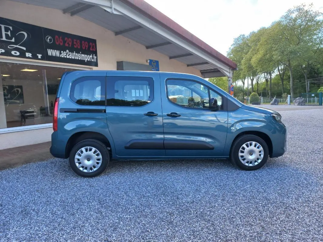 Vue de profil côté droit d'un Opel Combo Life Elegance M bleu 2025 avec protections latérales noires sur un sol gravillonné.