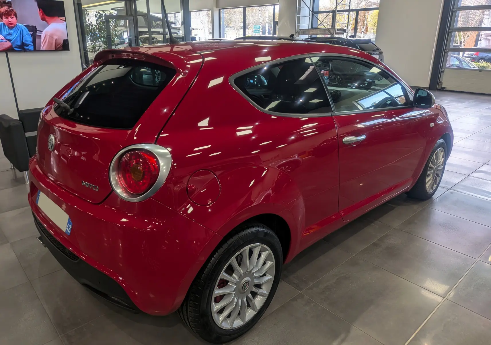 Vue 3/4 arrière droite d'une Alfa Romeo MiTo rouge Alfa 2018 dans un showroom lumineux.