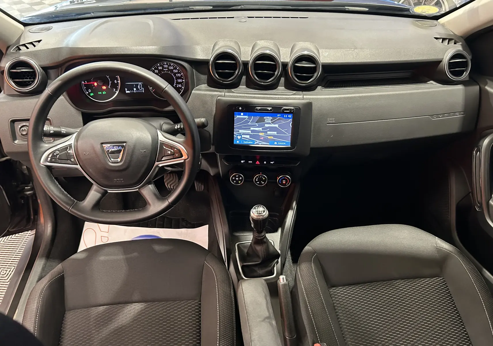 Vue intérieure avant du Dacia Duster noir 2018, avec tableau de bord, volant multifonction et écran GPS central.