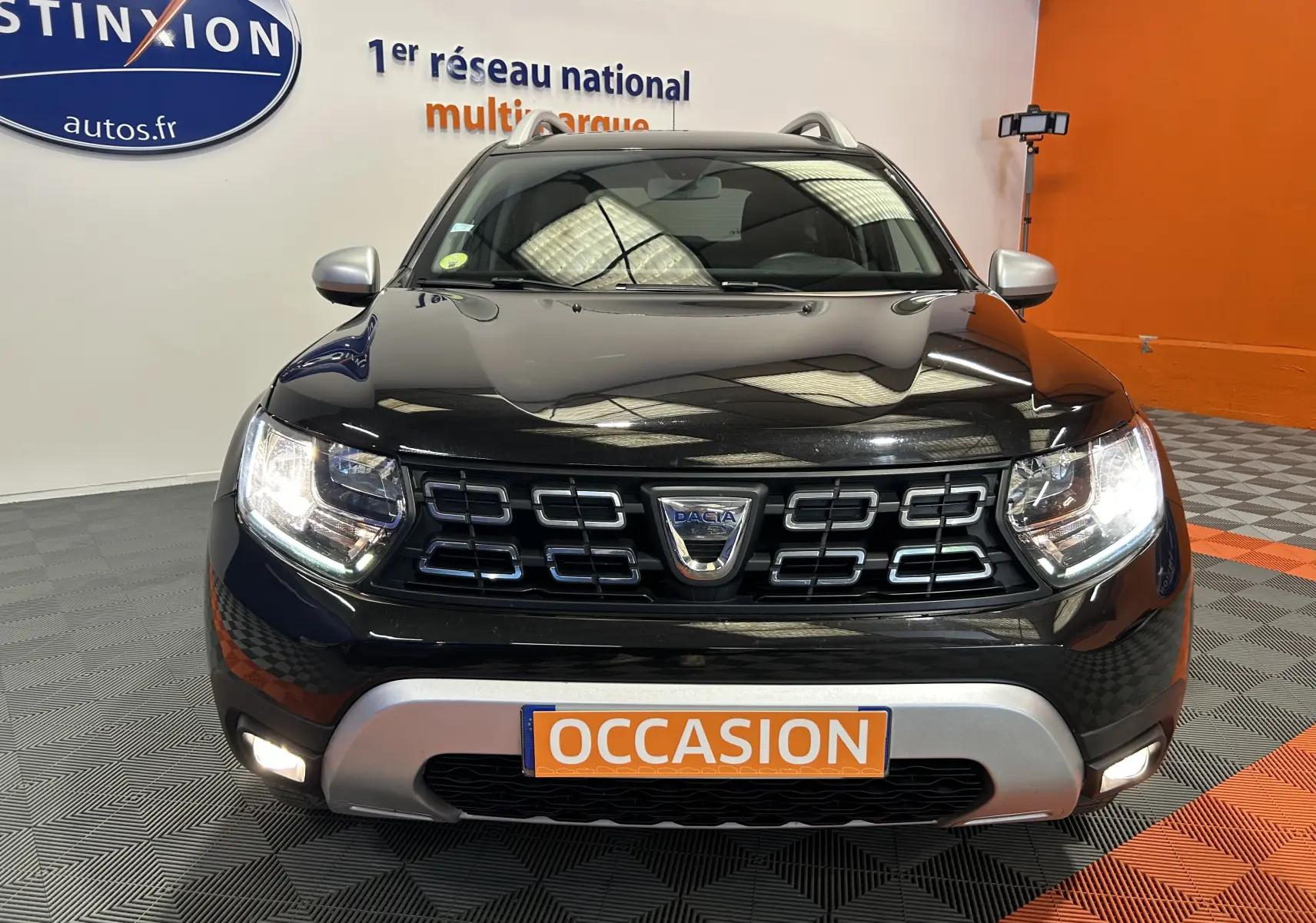 Vue frontale d'un Dacia Duster noir avec phares allumés et plaque "OCCASION" dans un showroom.
