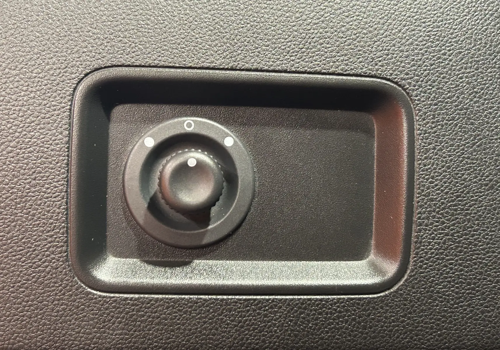Bouton rotatif noir encastré dans une surface texturée, utilisé pour régler les rétroviseurs d'un véhicule.