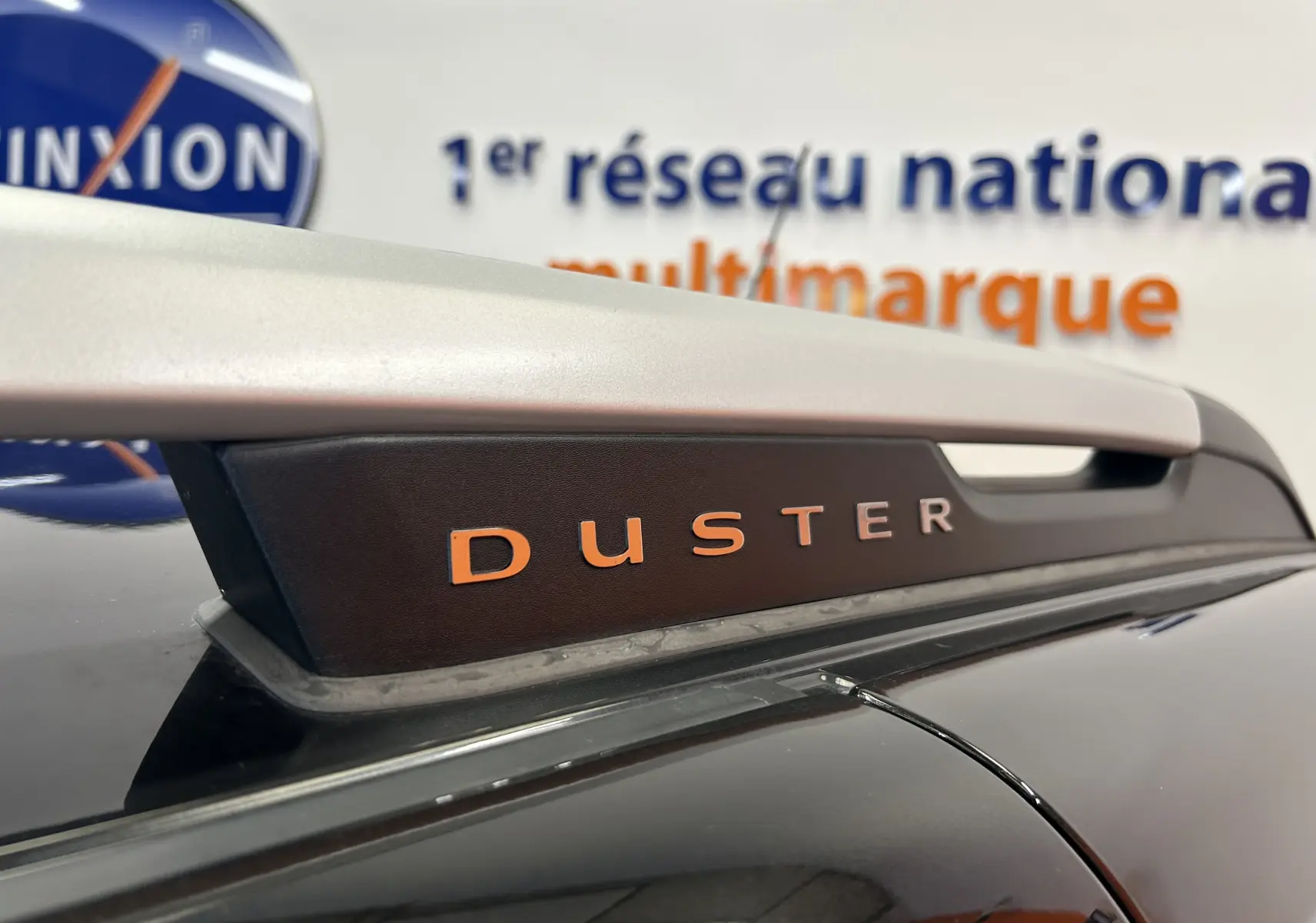 Gros plan sur la barre de toit noire avec logo DUSTER orange sur un Dacia Duster noir, arrière du véhicule visible.