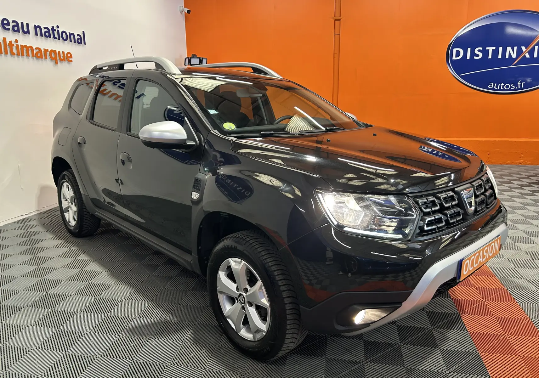 Vue 3/4 avant droite d'un Dacia Duster noir avec calandre chromée et barres de toit argentées en showroom.