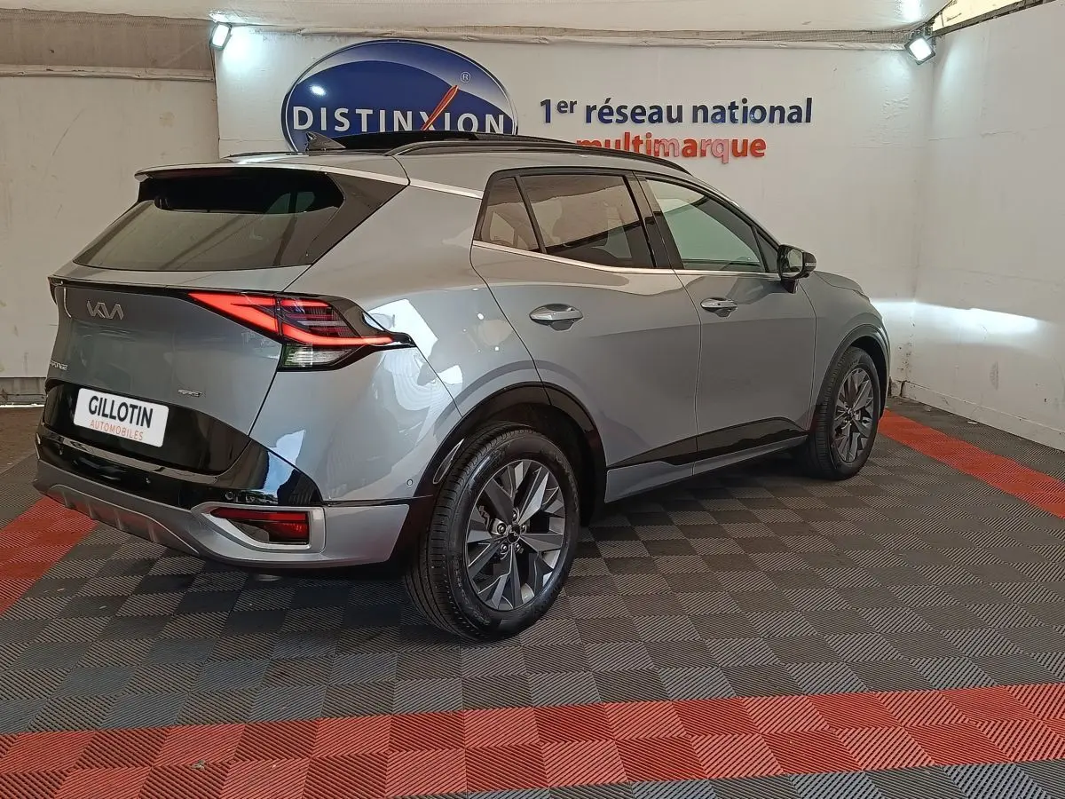 Vue 3/4 arrière droite d’un KIA Sportage gris clair avec feux arrière LED et jantes alliage foncé, en intérieur showroom.