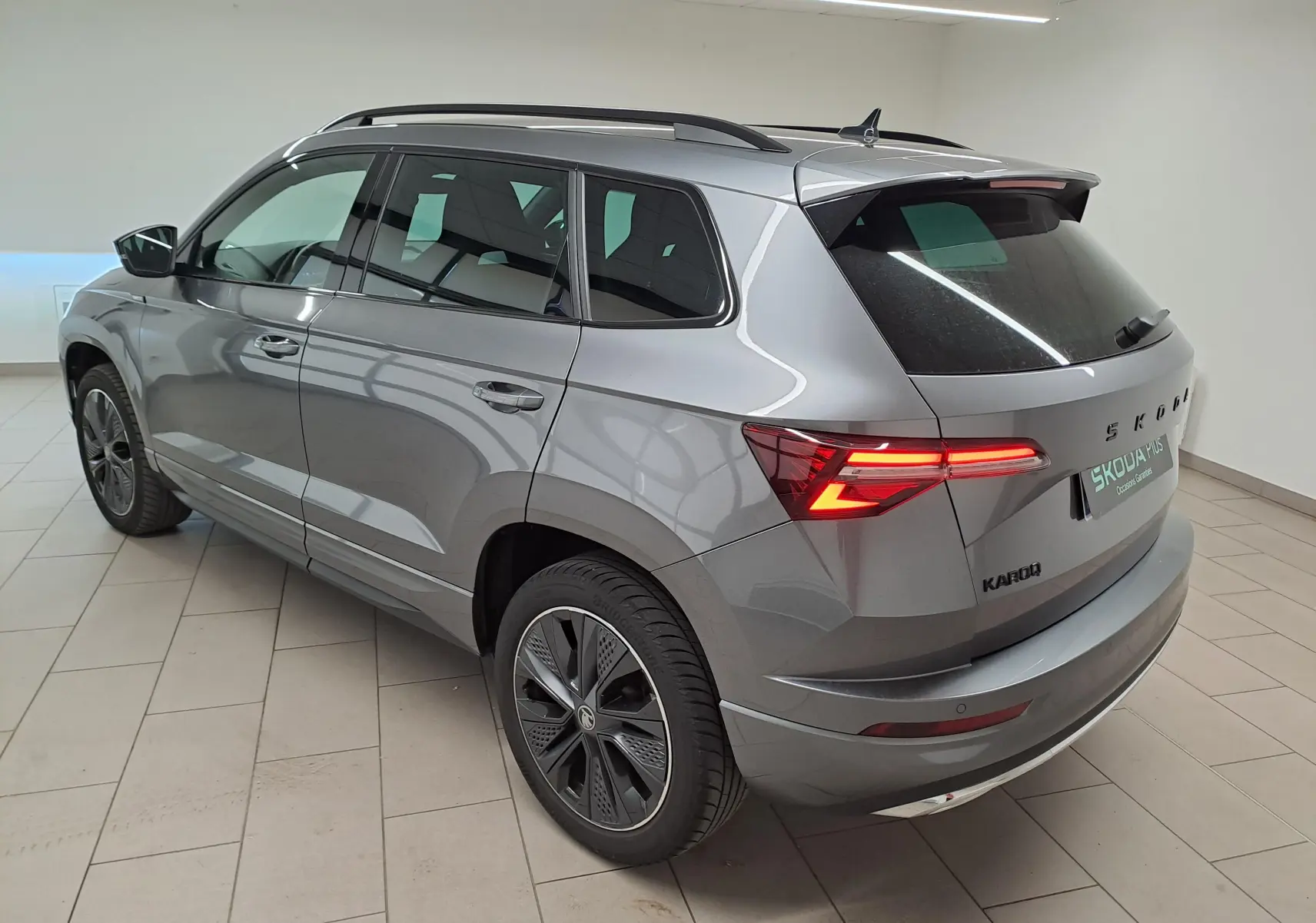 Vue 3/4 arrière droite d’un Skoda Karoq gris graphite 2025 avec feux allumés et hayon électrique en intérieur showroom.