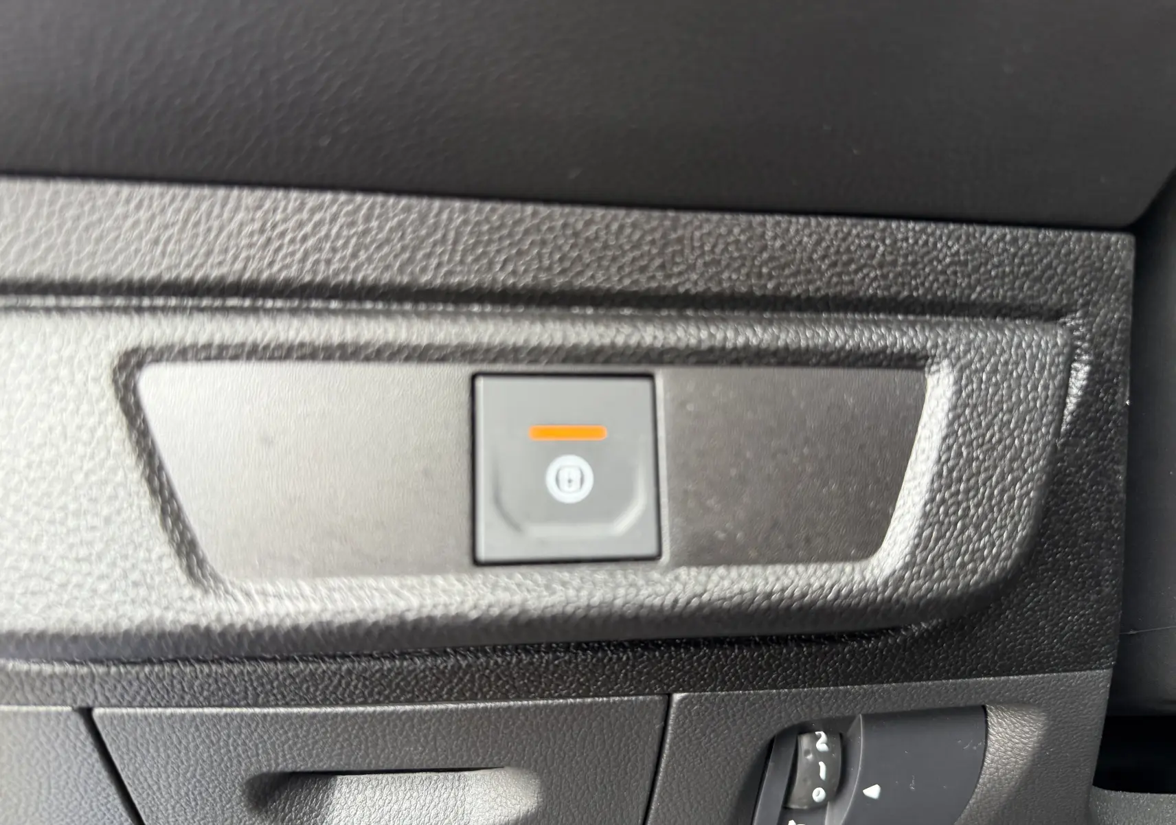 Gros plan sur le bouton de frein à main électrique et la molette de réglage des phares dans l’habitacle noir du Dacia Sandero Stepway bleu Iron.