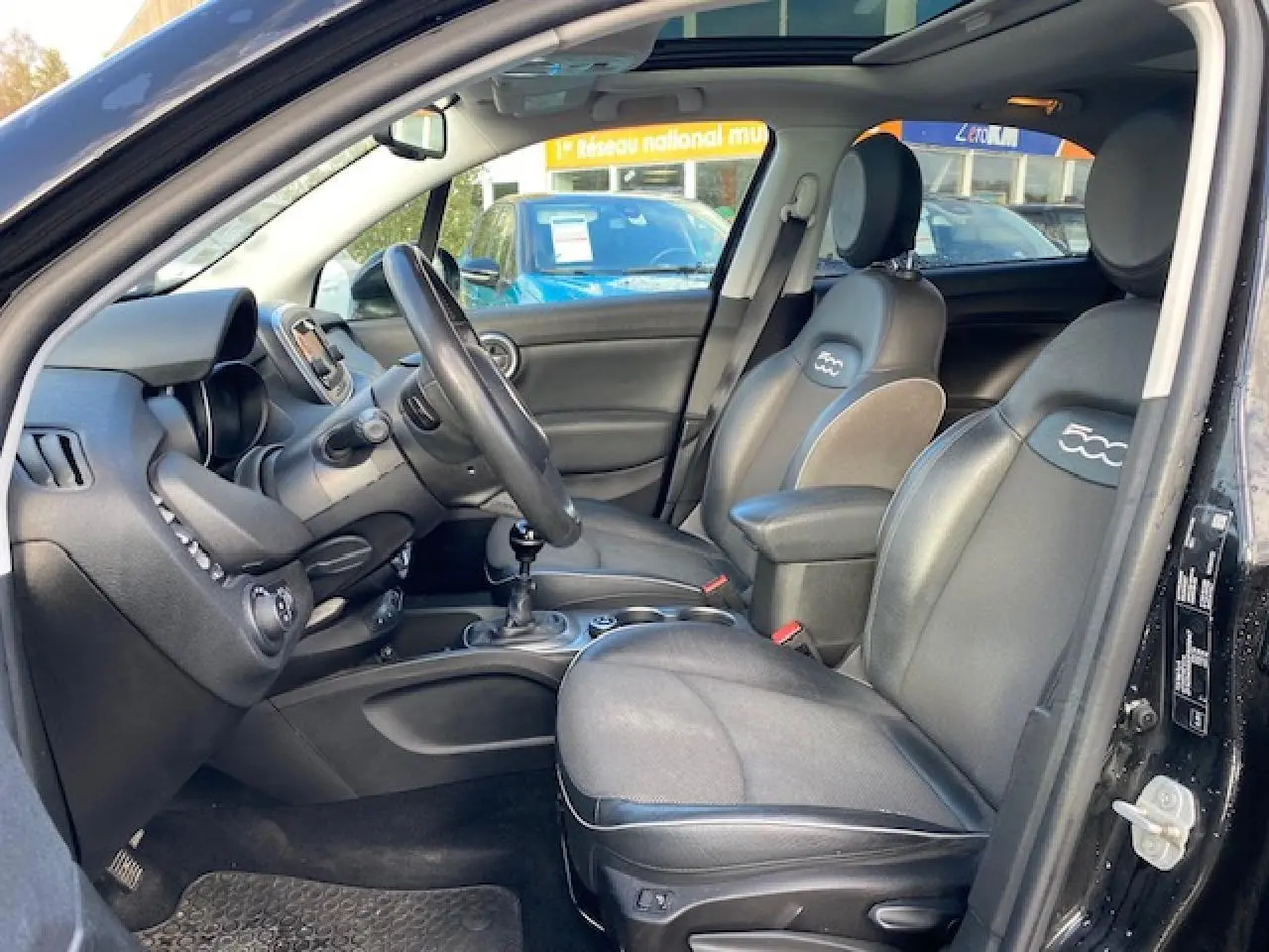 Intérieur noir vu côté conducteur d'une Fiat 500X 2016, sièges cuir avec logo 500 et toit panoramique ouvert.