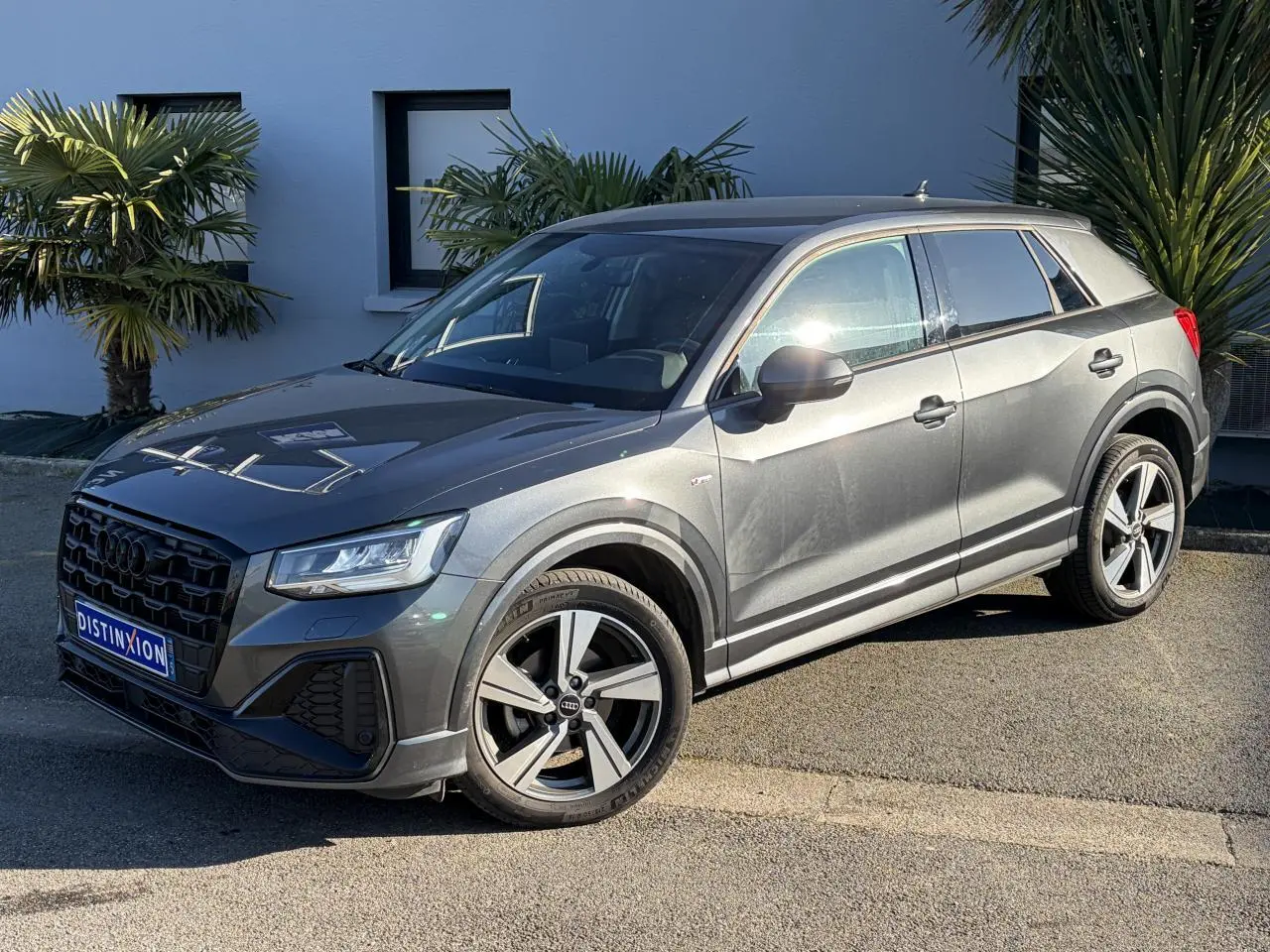 Audi Q2 35 TFSI S-Line gris Daytona en 3/4 avant droit, avec jantes alliage 18 pouces et vitres surteintées.