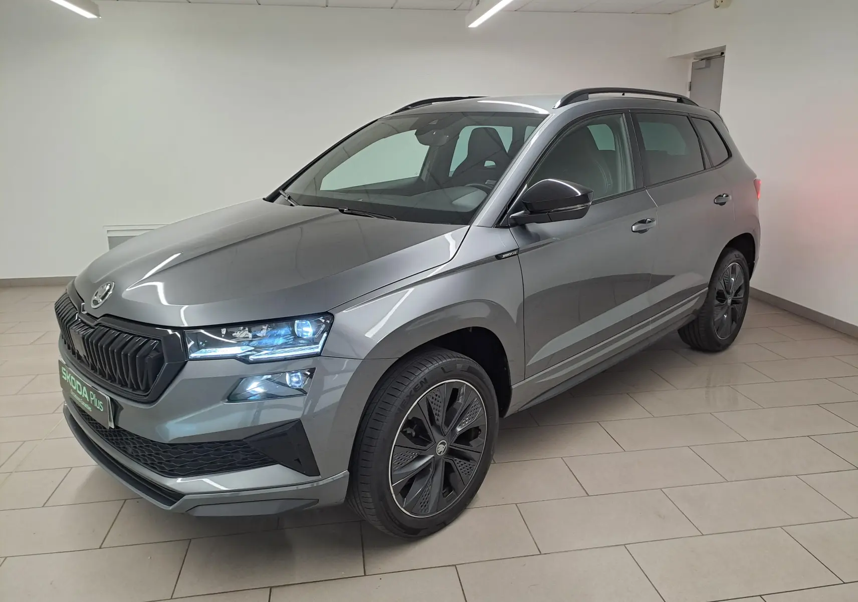 Vue 3/4 avant droite d’un Skoda Karoq gris graphite métallisé avec phares allumés et jantes noires sportline.