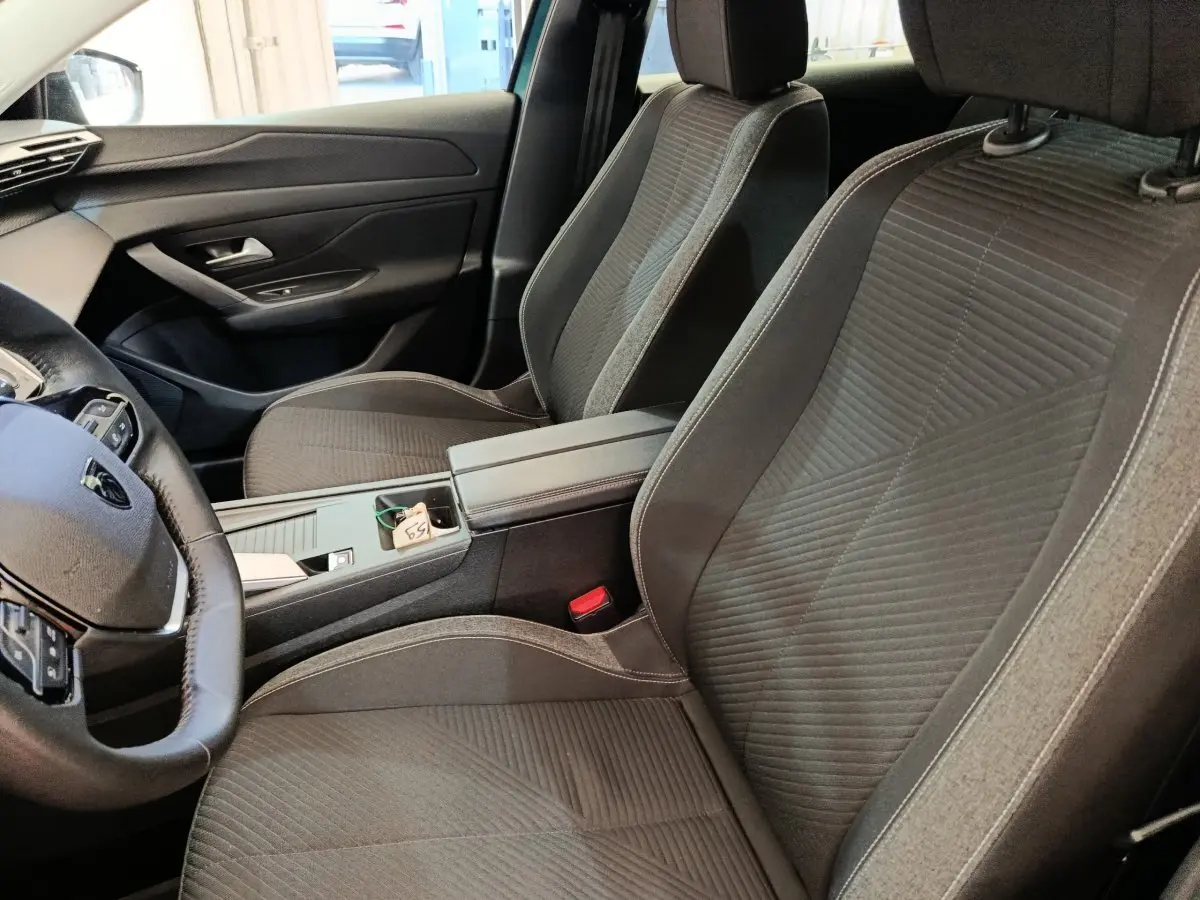Vue intérieure côté conducteur de la Peugeot 308 2023, sièges tissu gris et console centrale avec accoudoir.