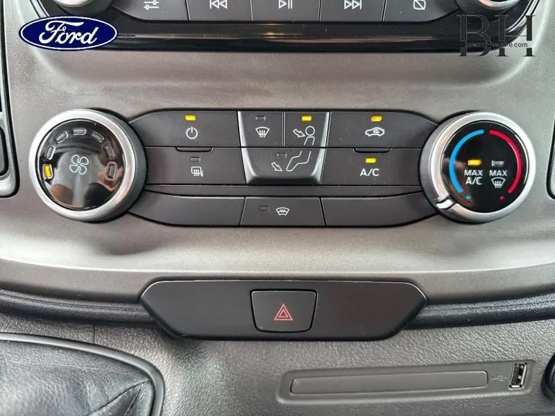 Vue rapprochée du panneau de climatisation du Ford Transit Custom blanc, avec commandes manuelles et bouton d'alerte au centre.