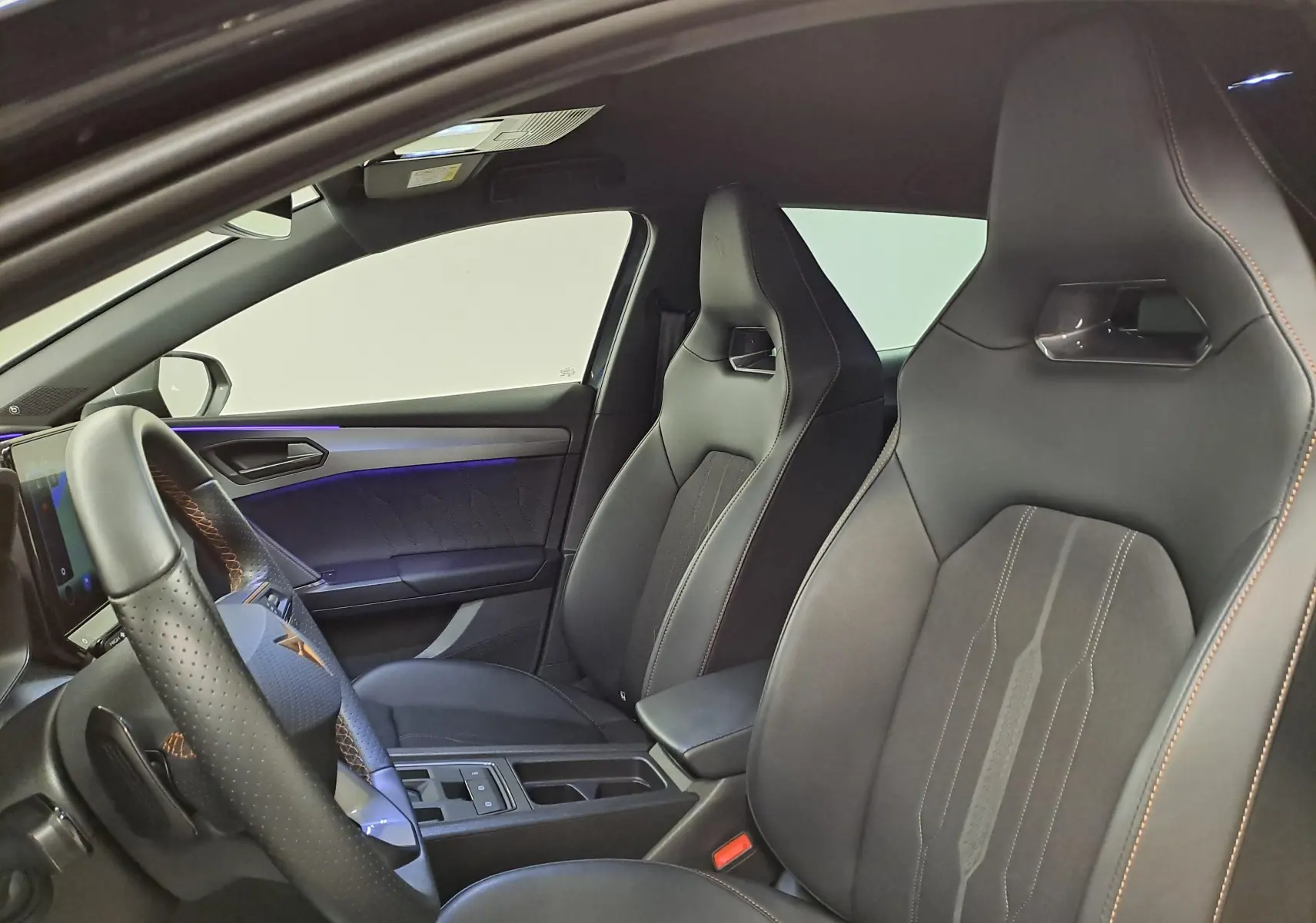 Intérieur avant droit de la CUPRA Leon 2023 gris magnétique, sièges sport noirs avec surpiqûres orange et volant cuir perforé.