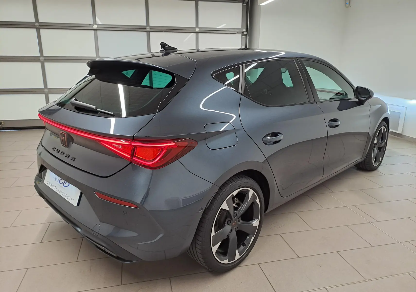 Vue 3/4 arrière côté gauche d'une CUPRA Leon 2023 gris magnétique avec feux arrière LED et jantes sport noires.