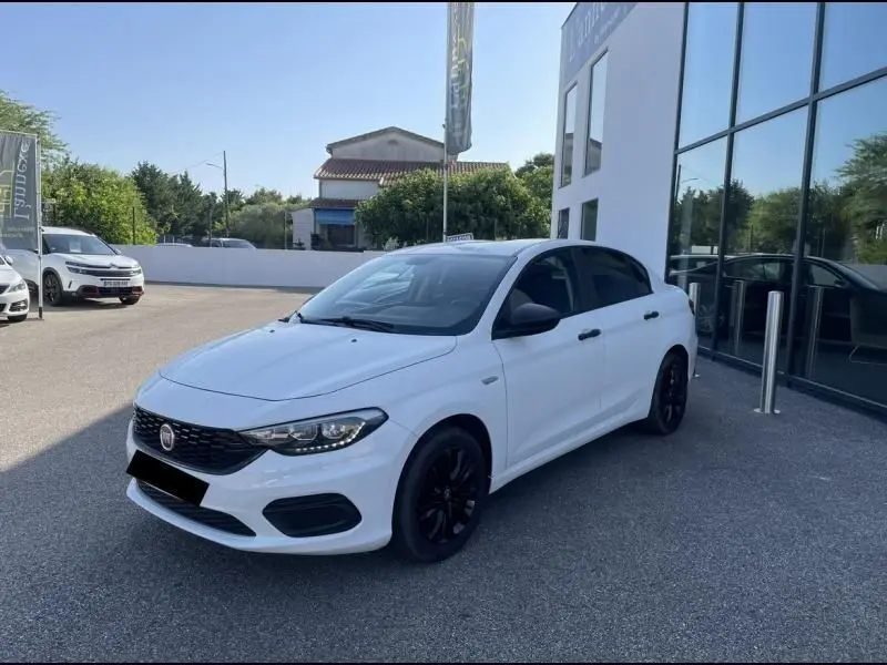 Vue 3/4 avant droite d'une Fiat Tipo blanche 2019 avec jantes noires, garée devant un bâtiment vitré.