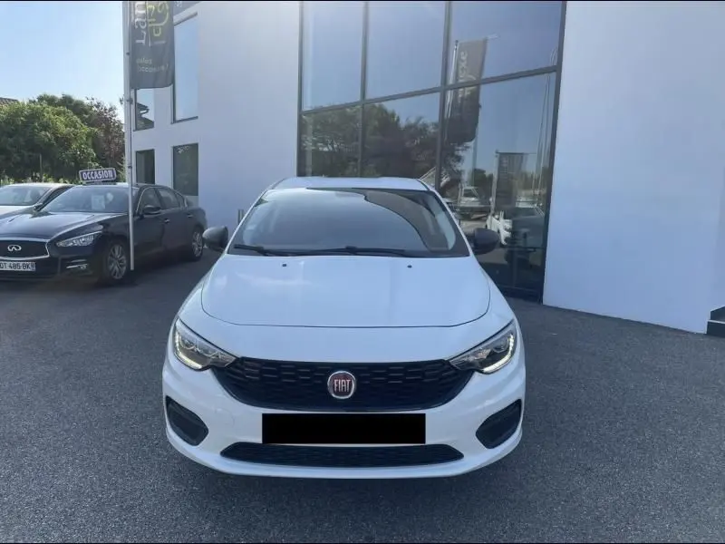 Vue frontale d'une FIAT TIPO blanche 2019 avec calandre noire et phares allumés, stationnée devant un bâtiment vitré.