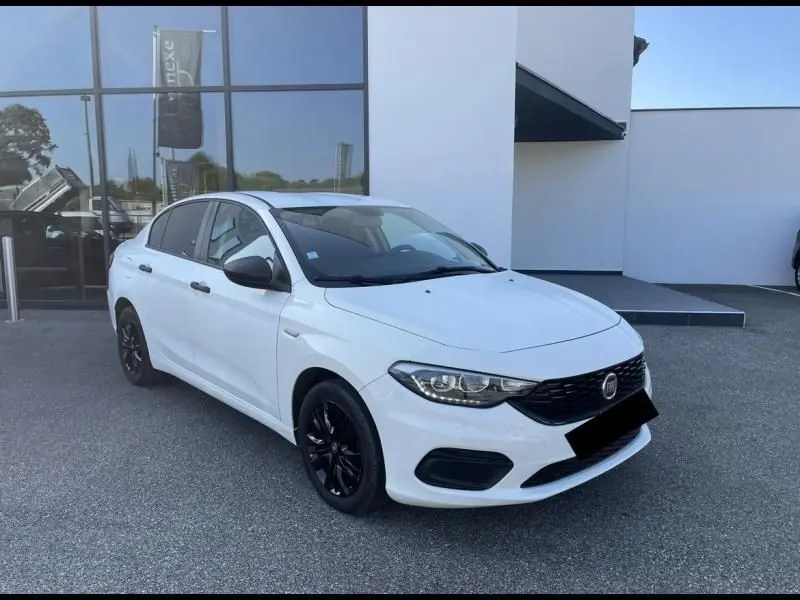 Vue 3/4 avant d'une Fiat Tipo blanche de 2019 avec jantes noires, garée devant un bâtiment moderne.