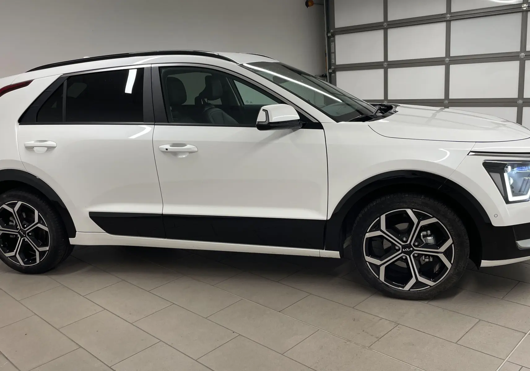 KIA NIRO hybride blanc vu de profil côté gauche, avec jantes noires et phares LED allumés dans un garage.