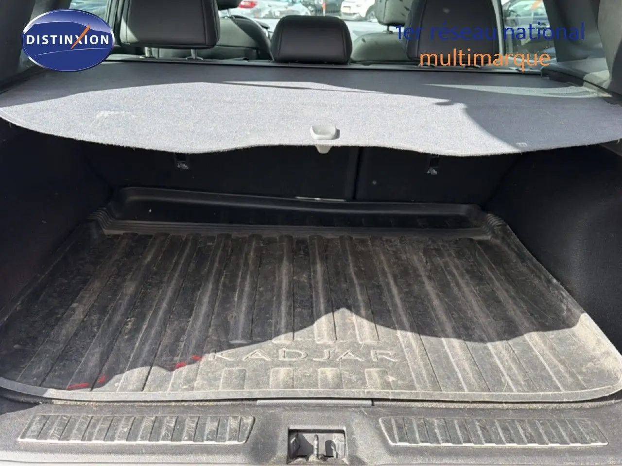 Coffre ouvert vu de l'arrière d'un Renault Kadjar gris Highland avec tapis de coffre marqué Kadjar et cache-bagages déployé.