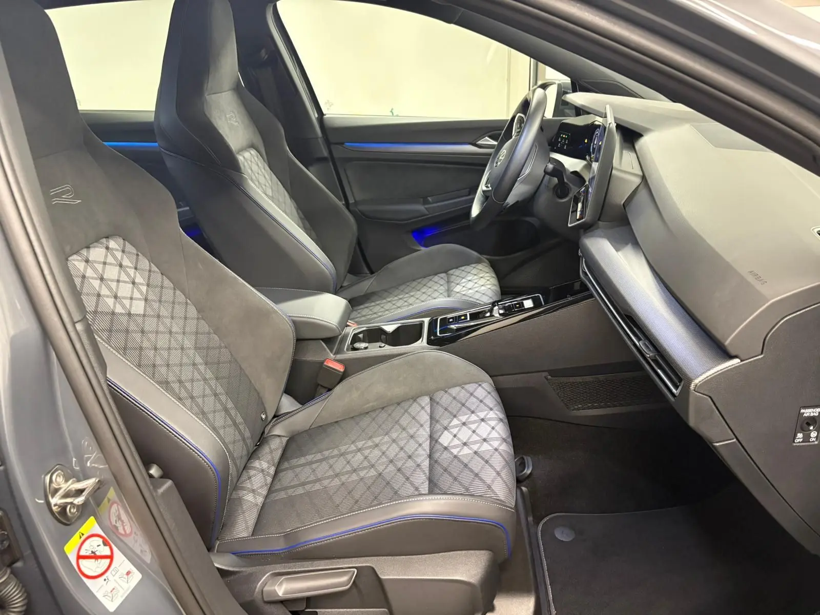 Vue intérieure côté gauche de la Volkswagen Golf 1.5 eTSI R-Line 2025 avec sièges sport gris à surpiqûres bleues et tableau de bord moderne.