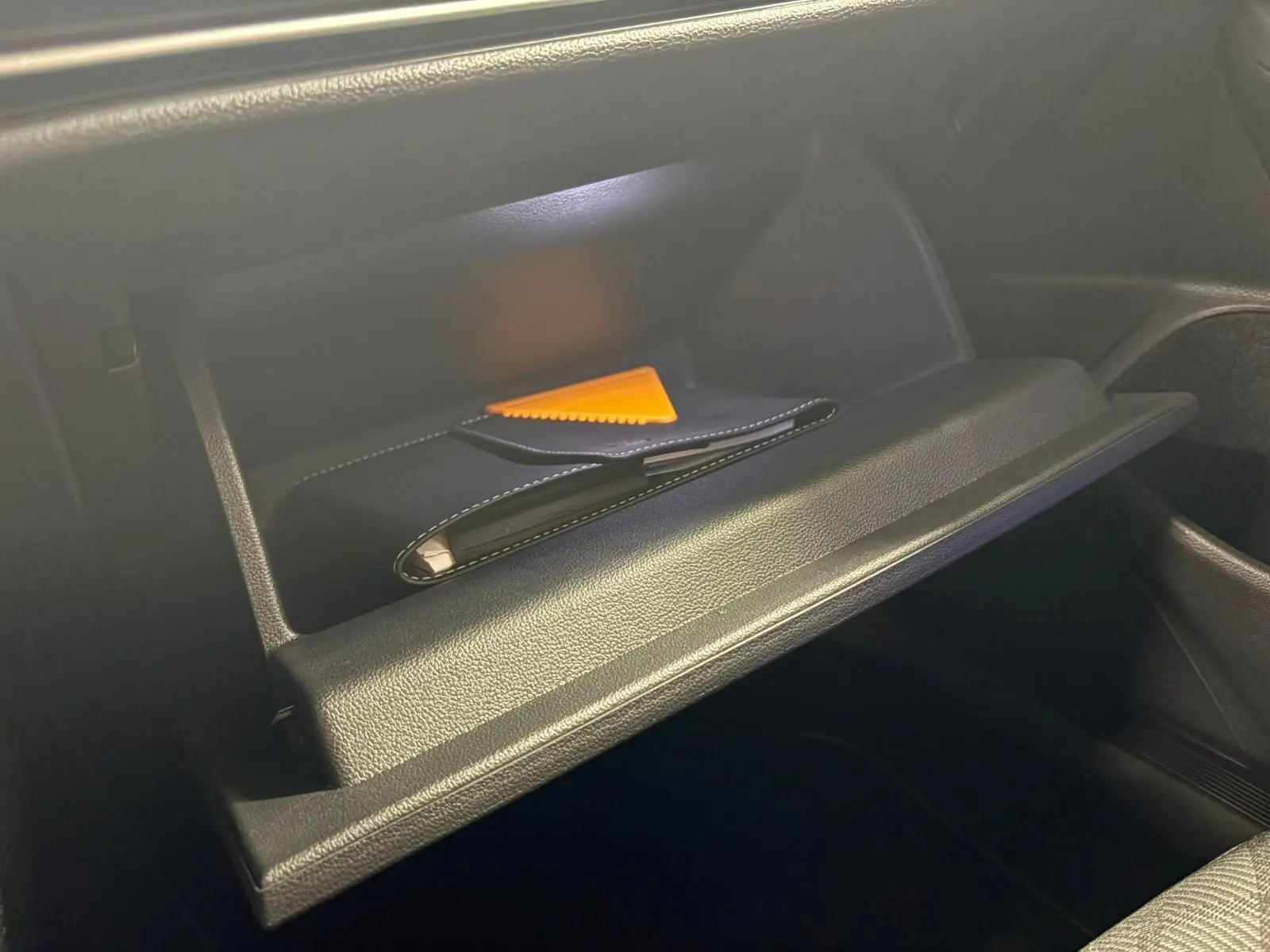 Gros plan sur la boîte à gants ouverte de la Volkswagen Golf 1.5 eTSI 150 DSG7 R-Line Edition avec un livret et un grattoir orange à l'intérieur.