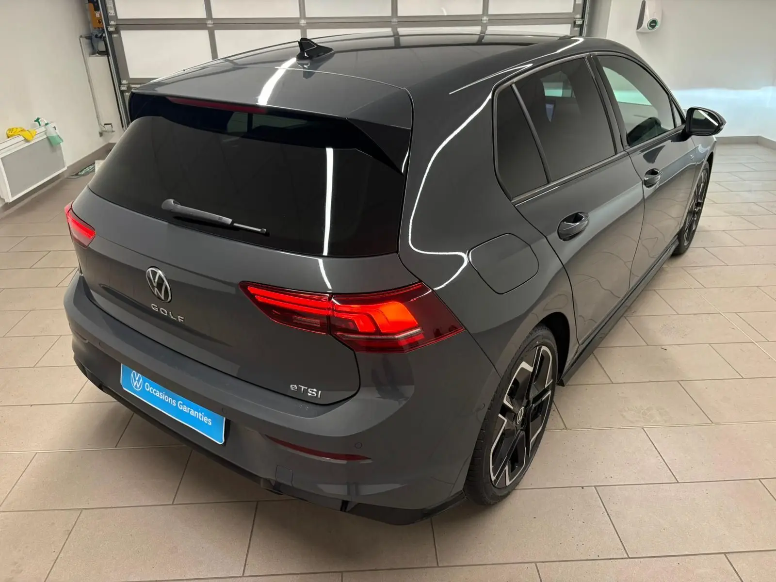 Vue 3/4 arrière droite d'une Volkswagen Golf 1.5 eTSI gris dauphin avec feux arrière allumés et jantes noires R-Line.
