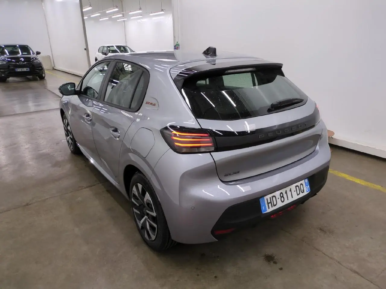 Vue 3/4 arrière droite d'une Peugeot 208 gris clair avec feux arrière à LED et bandeau noir brillant entre les feux.