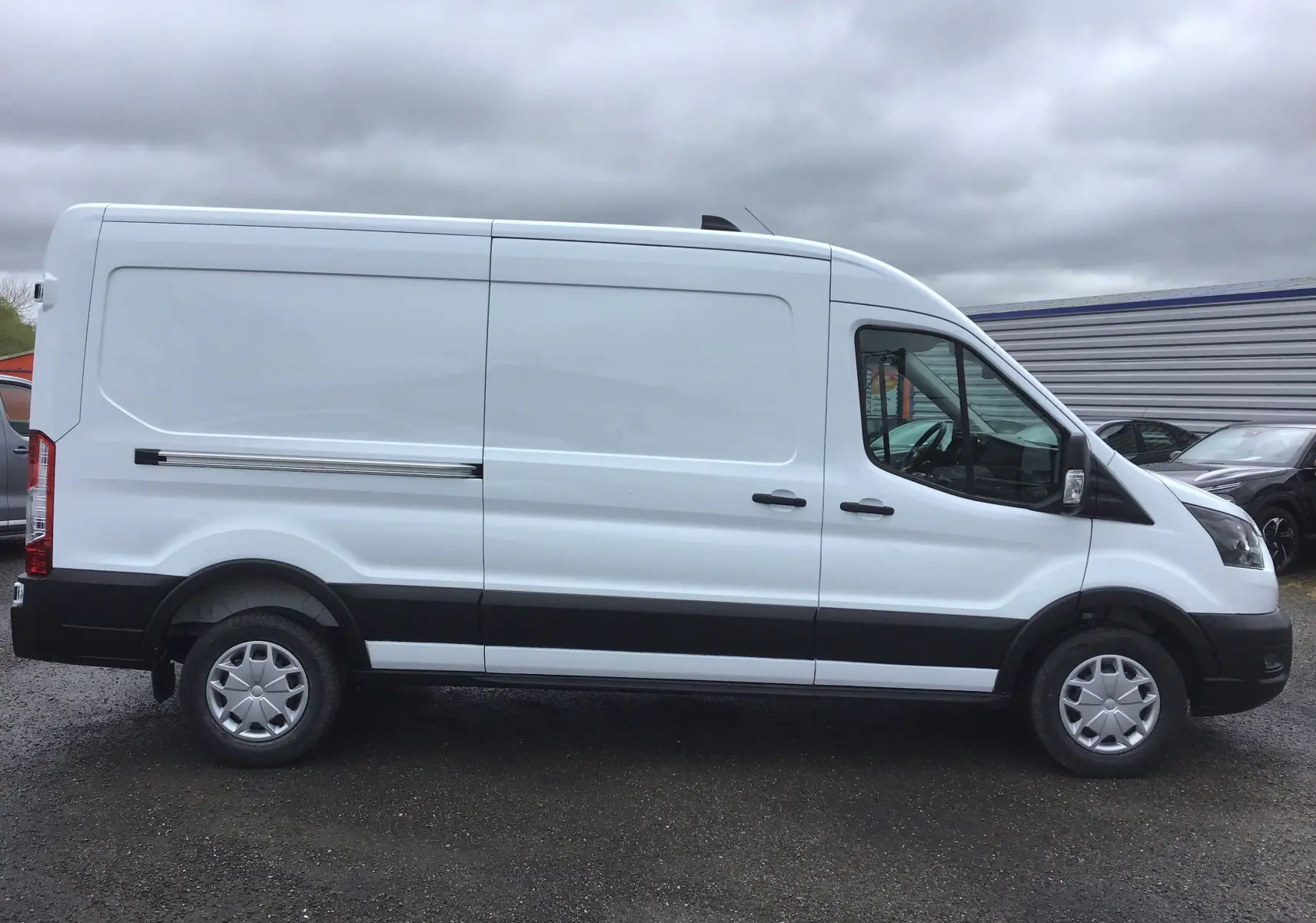 Vue latérale droite d'un fourgon Ford Transit blanc 2023 avec bandeau noir et jantes acier.