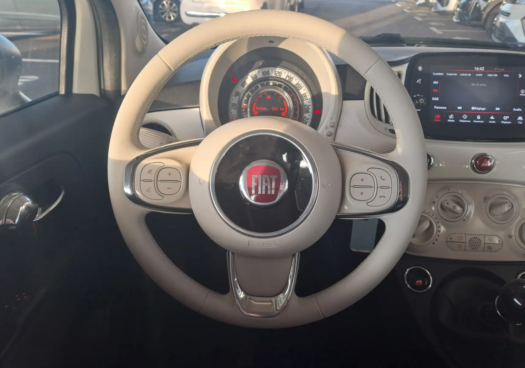 Vue intérieure centrée sur le volant beige de la Fiat 500 blanche 2023 avec tableau de bord et écran tactile visible.