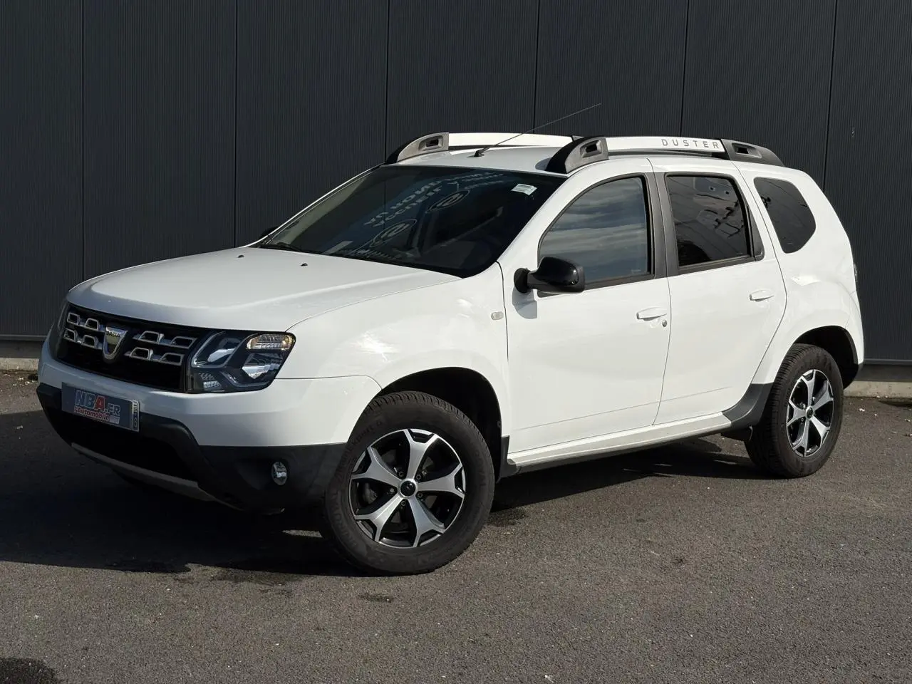 Dacia Duster blanc glacier en 3/4 avant droit avec barres de toit alu et jantes diamantées noires.