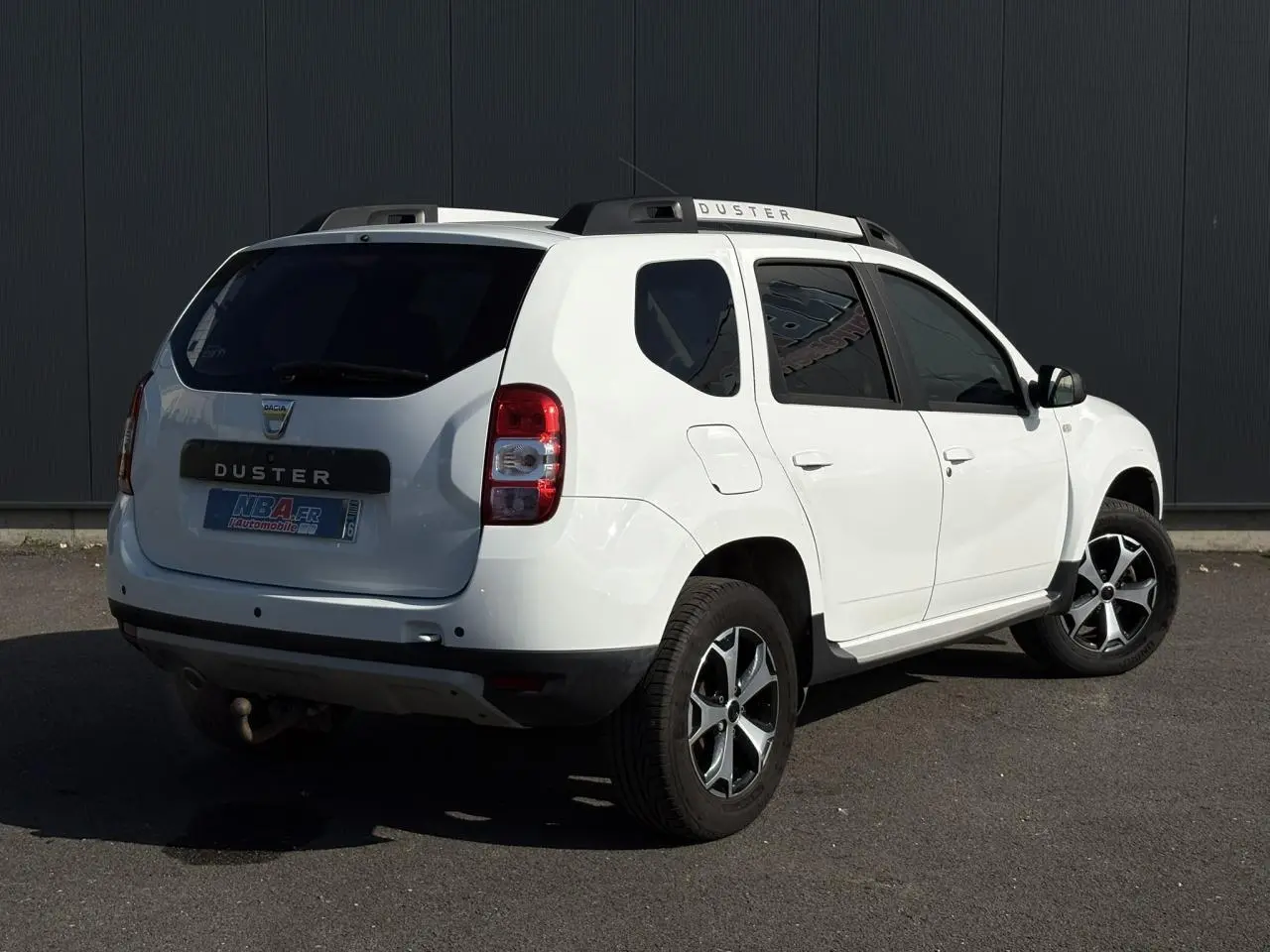 Dacia Duster blanc glacier vu en 3/4 arrière droit, avec barres de toit et attelage visible.