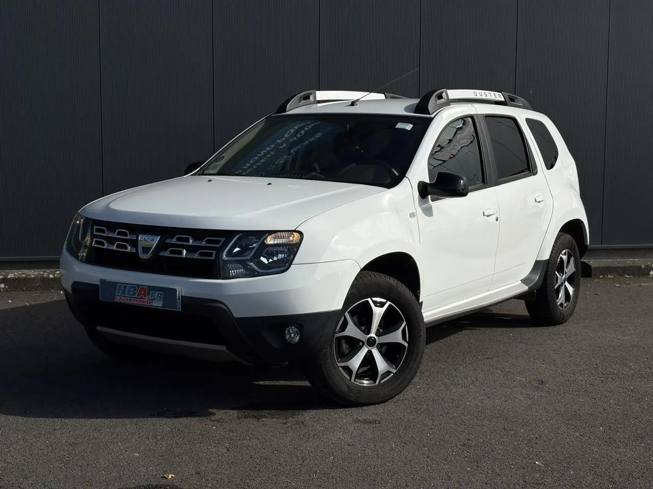 SUV Dacia Duster blanc glacier en 3/4 avant droit avec barres de toit alu et jantes diamantées noires.