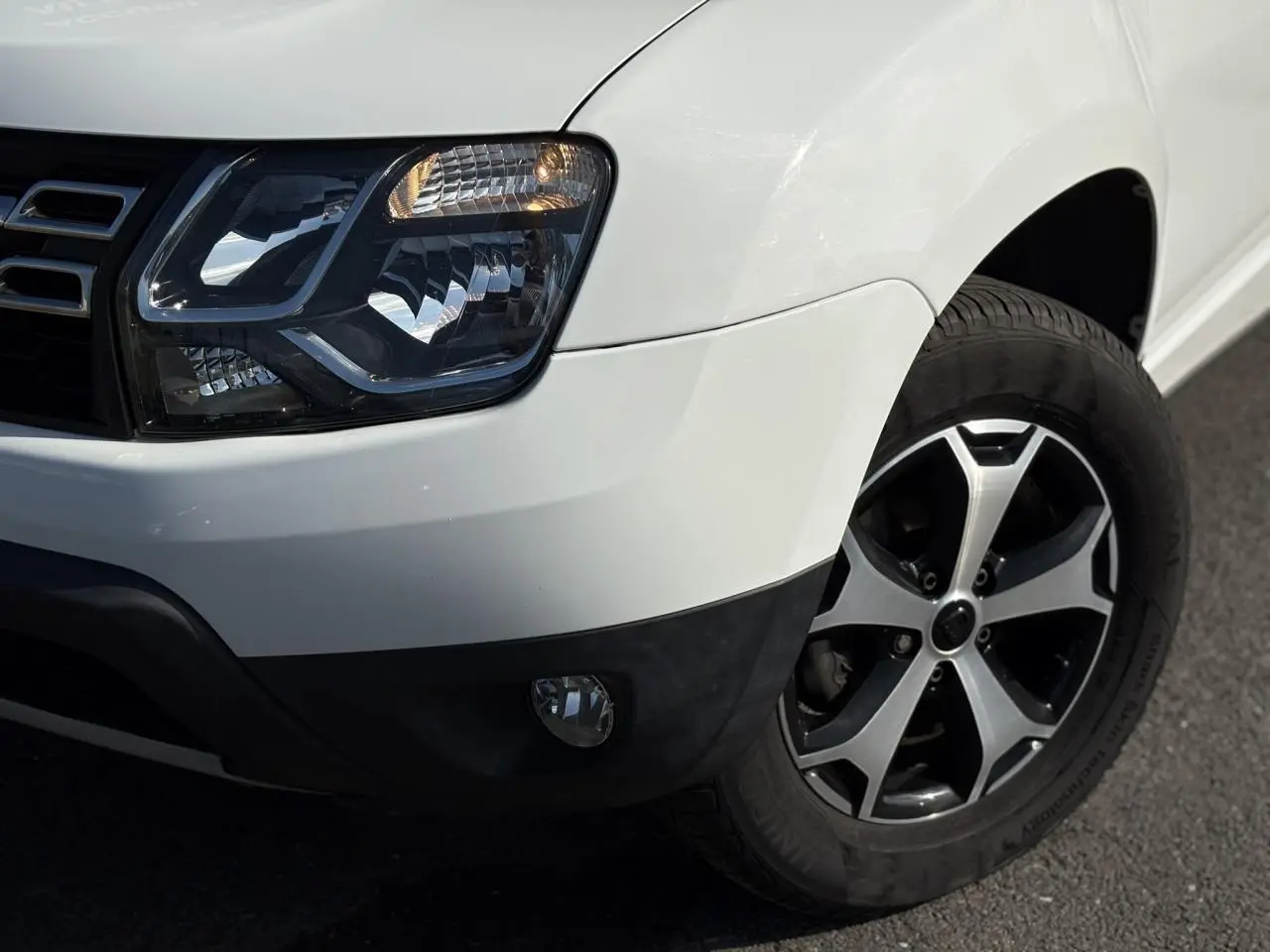 Gros plan sur l'avant droit du Dacia Duster blanc glacier, mettant en valeur le phare et la jante alu diamantée noire.