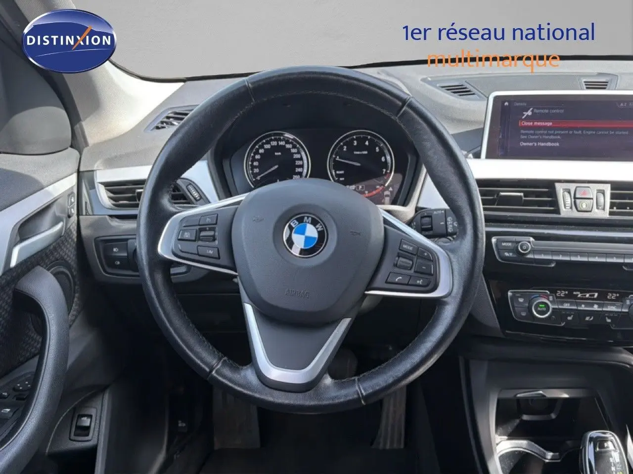 Vue intérieure centrée sur le volant noir d'une BMW X1 blanche, avec tableau de bord et écran multimédia visibles.