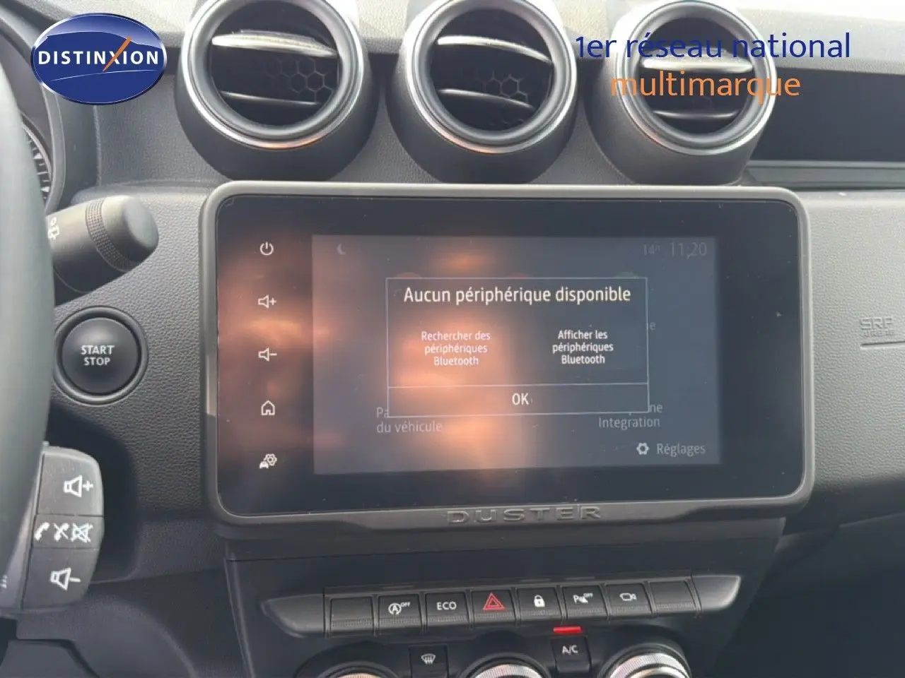 Vue rapprochée de l’écran tactile et de la console centrale du Dacia Duster bleu Iron, avec commandes et aérateurs ronds visibles.