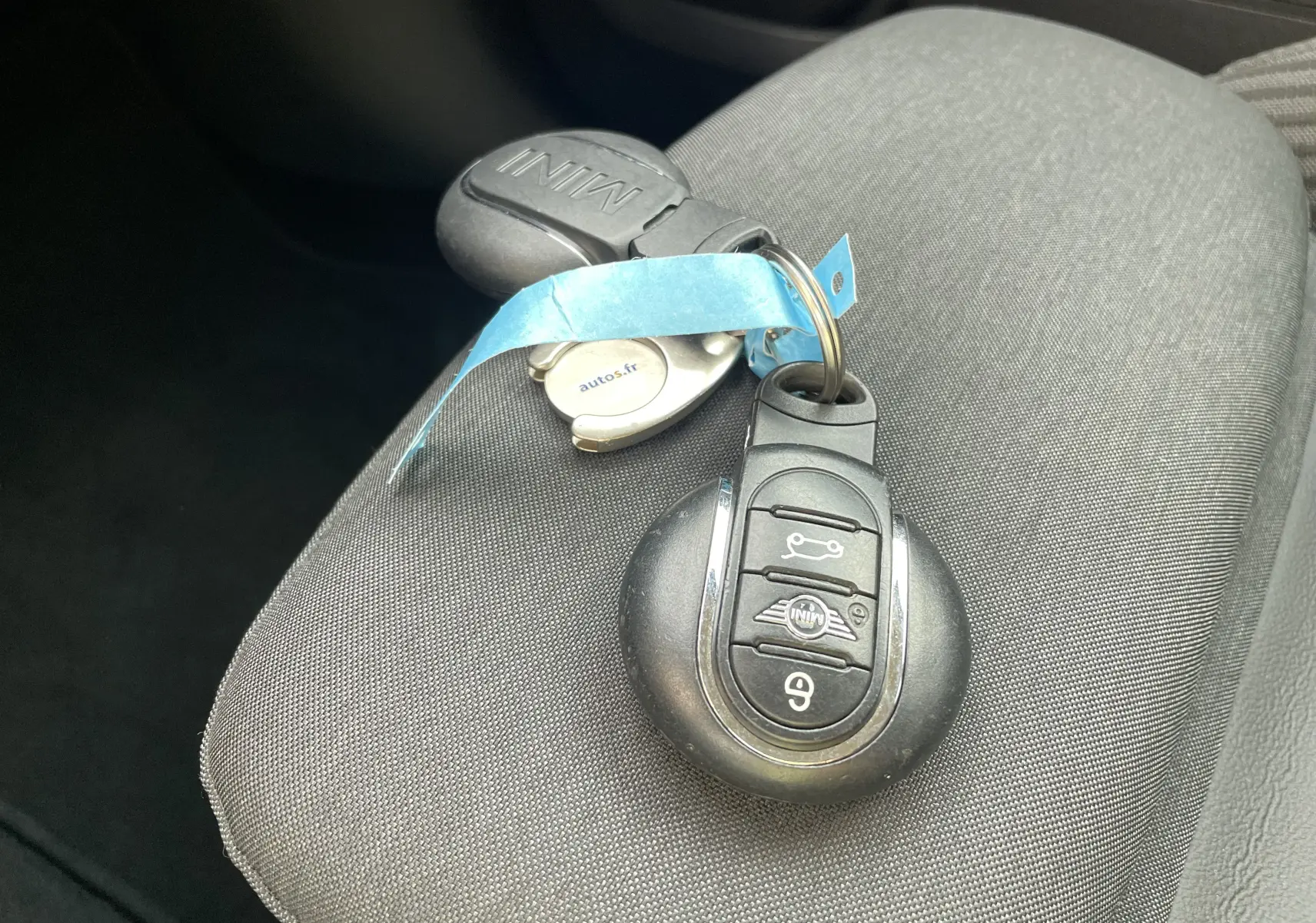 Gros plan sur la clé MINI posée sur un siège gris foncé, avec un porte-clé bleu et un badge autos.fr visible.