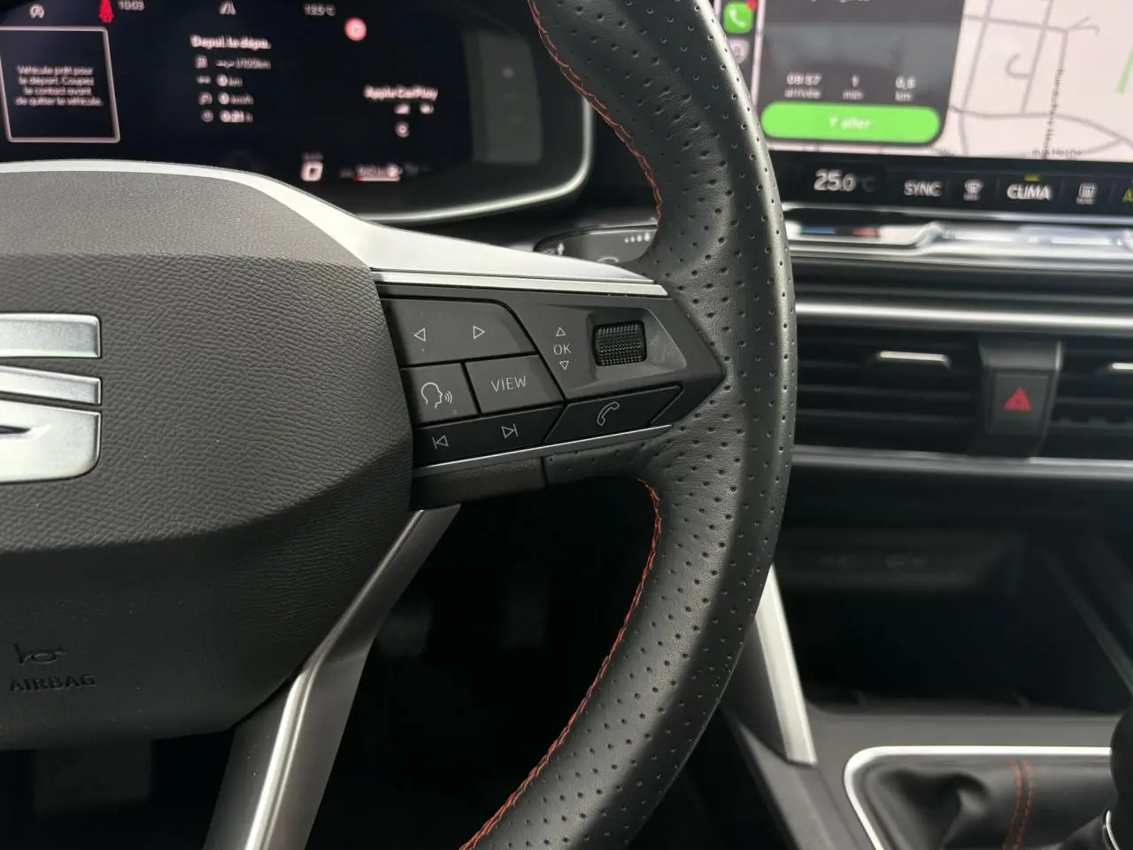 Gros plan sur le volant en cuir noir à surpiqûres rouges du SEAT Leon 2025, avec commandes multifonctions et écran tactile en arrière-plan.
