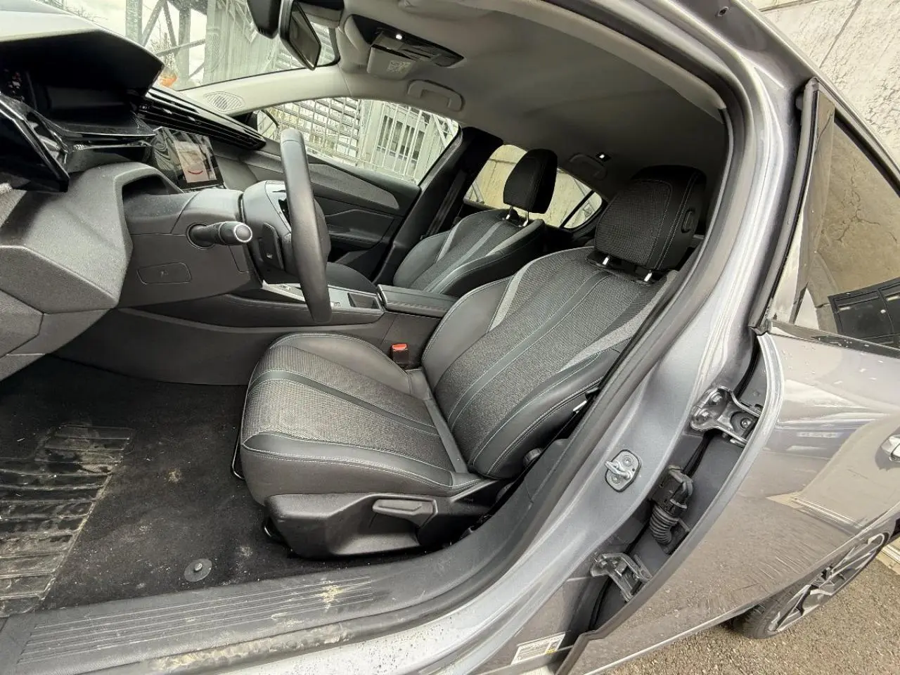 Intérieur de la Peugeot 308 1,2 Hybrid 2025 vu côté conducteur, sièges tissu et cuir gris, tableau de bord moderne.
