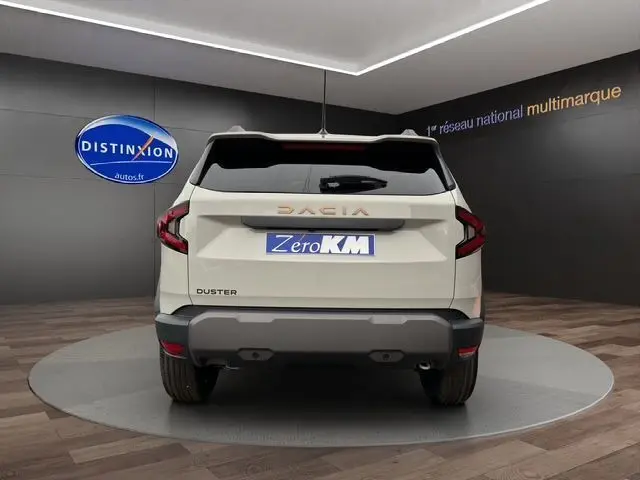 Vue arrière d'un Dacia Duster blanc avec logo et feux LED, stationné en showroom sur fond sombre.