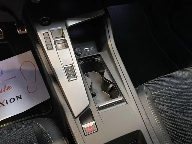 Vue rapprochée de la console centrale noire de la Peugeot 308 Hybrid 2026, avec commandes et porte-gobelets visibles.