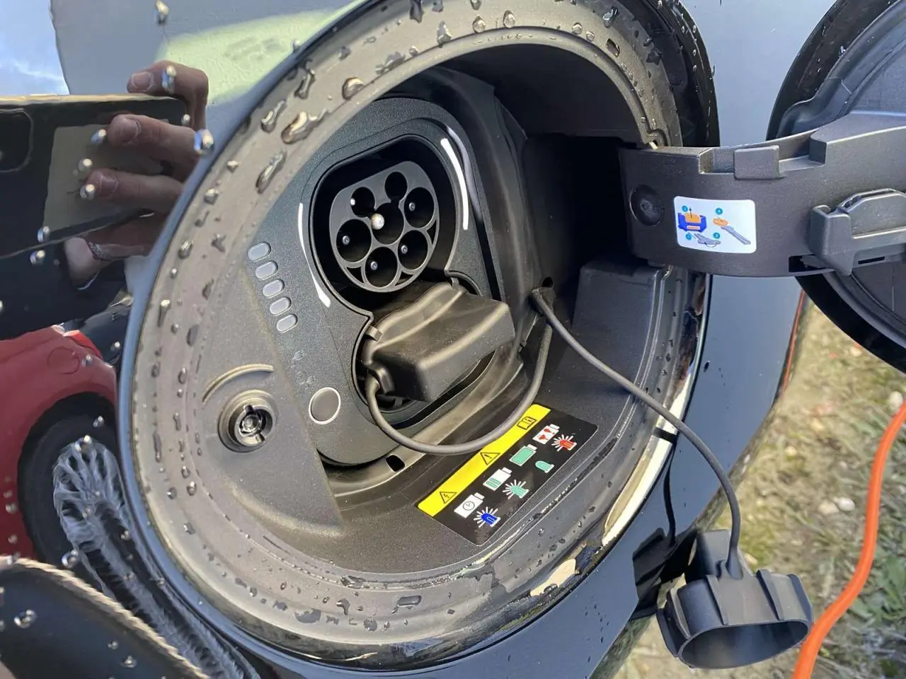 Prise de recharge électrique ouverte sur une FIAT 500 noire, avec câble de charge visible et gouttes d'eau.
