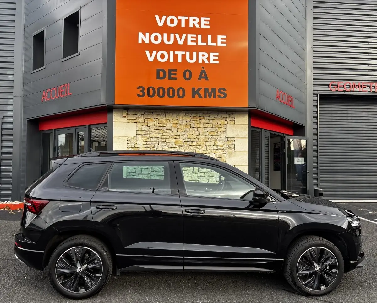 Profil côté gauche d'un Skoda Karoq noir 2025 garé devant un bâtiment avec panneau orange "Votre nouvelle voiture".