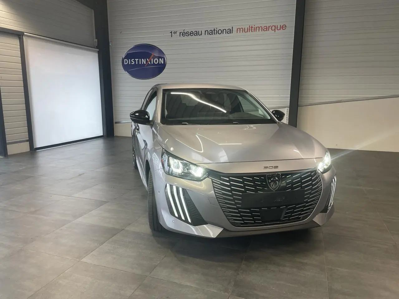 Peugeot 208 gris Artense en 3/4 avant droit, avec calandre noire et feux LED allumés dans un showroom.
