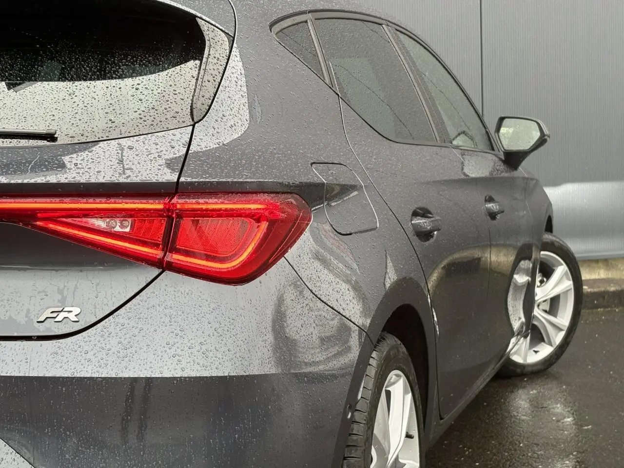 Vue 3/4 arrière droite d'une SEAT LEON 1.5 e-TSI FR gris magnétique avec feux arrière LED allumés sous la pluie.