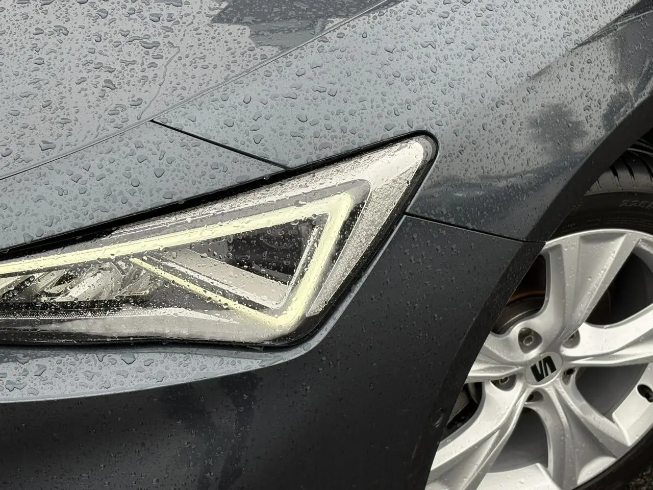 Gros plan sur le phare avant gauche et la jante alliage 17'' d'une SEAT LEON Gris Magnétique sous la pluie.