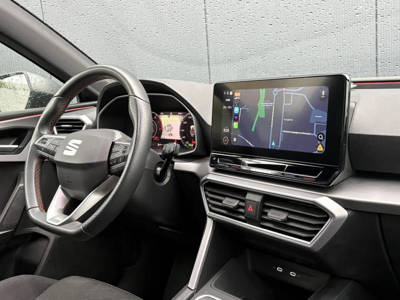 Vue intérieure côté conducteur du tableau de bord du SEAT Leon 2024 avec écran tactile et volant cuir noir à surpiqûres rouges.